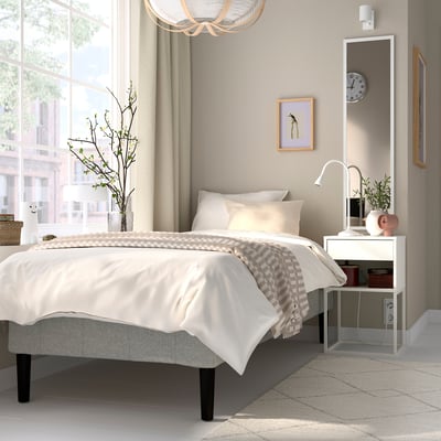 Minimalistiskt sovrum med vit säng, spegel, nattduksbord, lampa, växter, stora fönster.