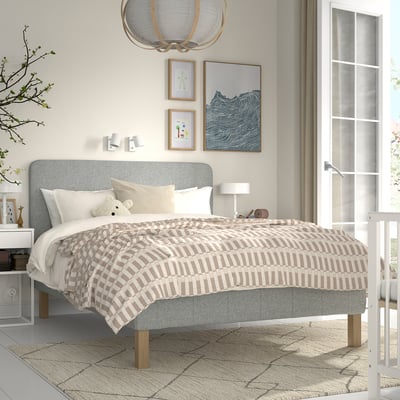 Modern grå säng med randigt beige täcke, vita kuddar. Stor konst, björnleksak, träben. Storlek: kung.