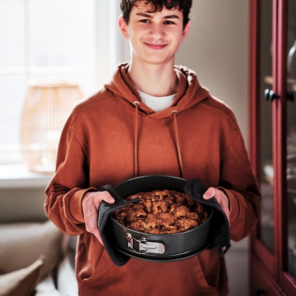 Tonåring i brun hoodie håller svart panna med bakad mat, troligen tas bort från ugnen med hjälp av ugnsvante.