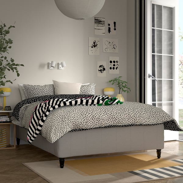 Sovrum med modern minimalistisk stil. Grå säng i mitten med prickig och randig säng. Väggkonst, gul lampa och växter. Stort fönster till höger.