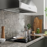 Modernt kök med elegant fläkt, stänkskydd i marmor, induktionshäll, stekpanna med ånga, träredskap, burkar och växter.