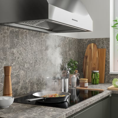 Modernt kök med elegant fläkt, stänkskydd i marmor, induktionshäll, stekpanna med ånga, träredskap, burkar och växter.