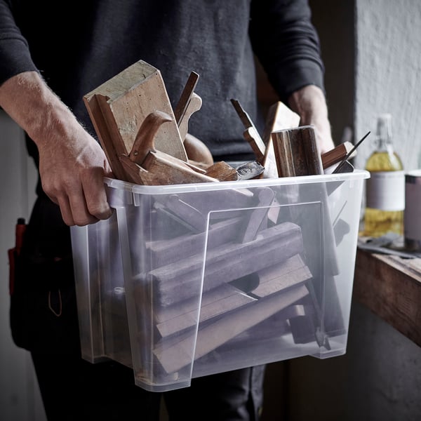 En person håller i en genomskinlig plastbehållare fylld med träverktyg, redo att organiseras och förvaras.