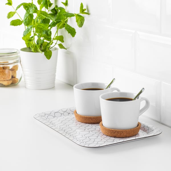 Två vita kaffekoppar på korkunderlägg står på en mönstrad bricka med vinklade kanter, placerade på en vit bänkskiva med en grön växt och kakor i närheten.