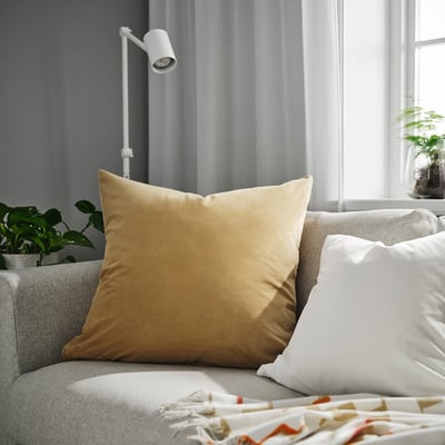 Stor guldfärgad SANELA kudde på beige soffa bredvid vit kudde, modern lampa, grön växt och säng med färgrik filt.