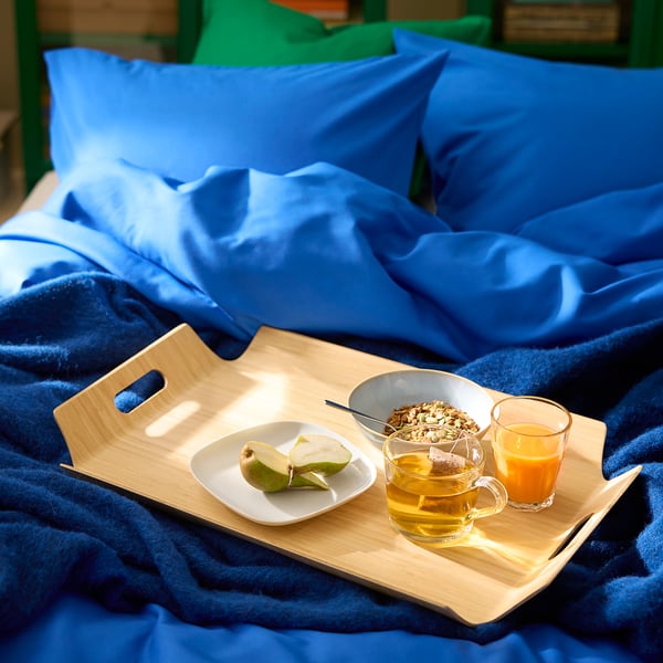 Frukostbricka av bambu på blå säng med frukt, granola, te och juice.