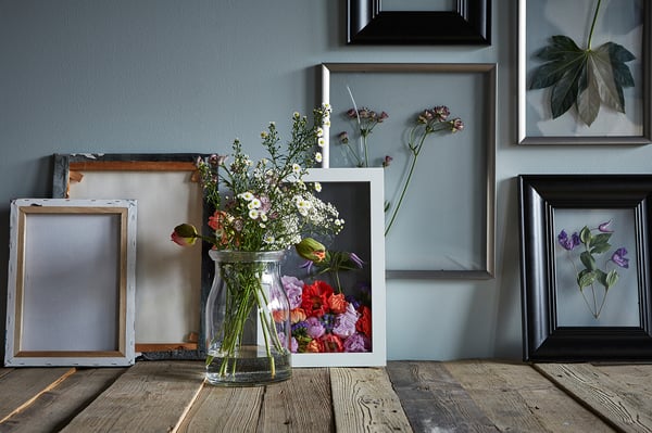 En väggdisplay med flera bildramar i olika färger och storlekar, inklusive en framträdande vit ram med ett blommigt arrangemang.