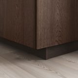 Skåp i mörkt trä med synlig ådring, massiv ekkant på sockeln, står på ett ljust trägolv och visar upp SINARP-fronternas elegans och detaljer.