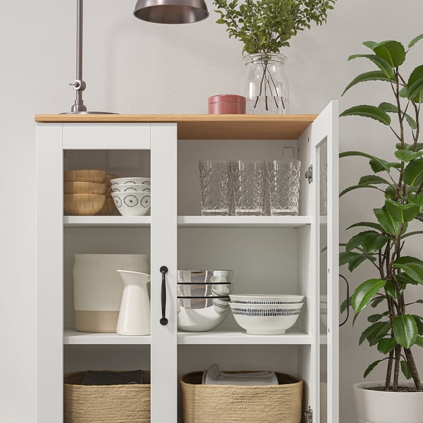 Vitt skåp med glasdörrar som visar skålar, glas och korgar. Översta hyllan har växt, burk och vas. Snygga handtag, justerbara hyllor.