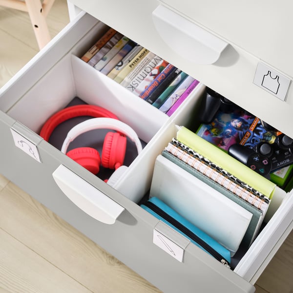 Vit byrå SMÅSTAD med lådor. Innehåller röda hörlurar, böcker, bärbara datorer och speltillbehör. Organiserad, barnvänlig design.