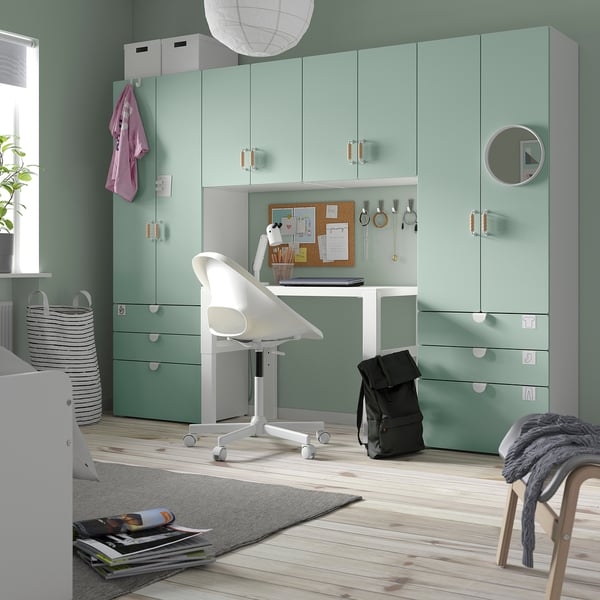 Ljust hemmakontor: ljusgröna SMÅSTAD-enheter med vitt skrivbord och stol, svart ryggsäck, grå filt, naturligt trägolv, mjuk matta och växt.