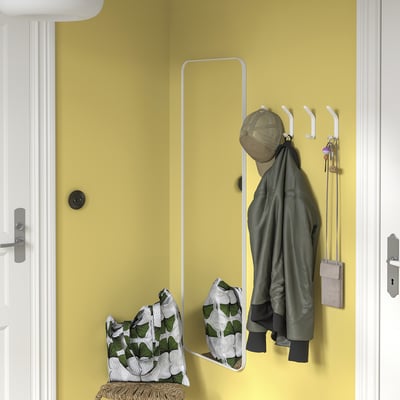 Gul vägg med vita SNÖRAKA-krokar som håller grön jacka, väska och nycklar. Spegel nedan, korgbänk med kudde.