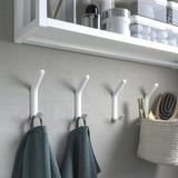 Vita dubbelkrokar på väggen med handdukar hängande, hylla ovanför med badrumsartiklar.