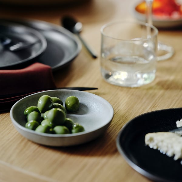 En liten skål med ljusgröna ärtor på en grå tallrik, ett glas med klart vatten och ett litet fat med vit, smulig mat bredvid dem på ett träbord.