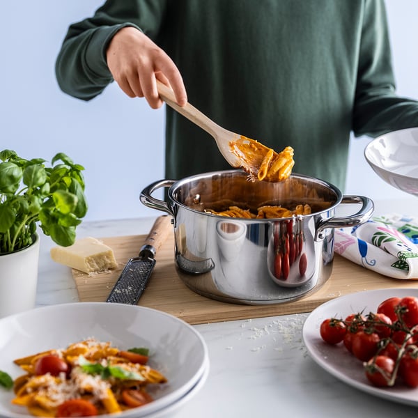 Person som rör pasta i kastrull, serverar med träslev. Innehåller ost, tomater, basilika och rivjärn.