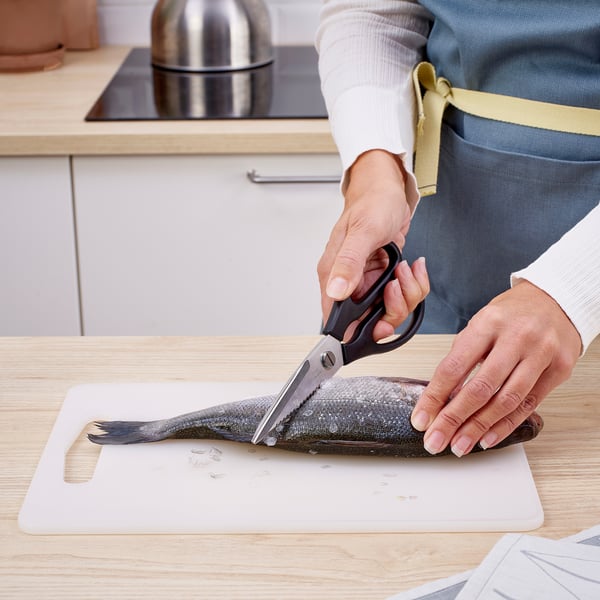 Personen använder sax för att klippa fisk på en vit skärbräda i köket.