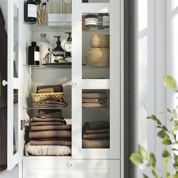 Vitt badrumsskåp med glasdörrar som visar vikta handdukar och toalettartiklar, som erbjuder förvaring och organisation med en klassisk design.