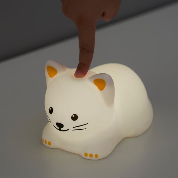 En hand rör vid en TÖVÄDER nattlampa formad som en vit katt med orange fläckar och öron och avger ett mjukt sken.