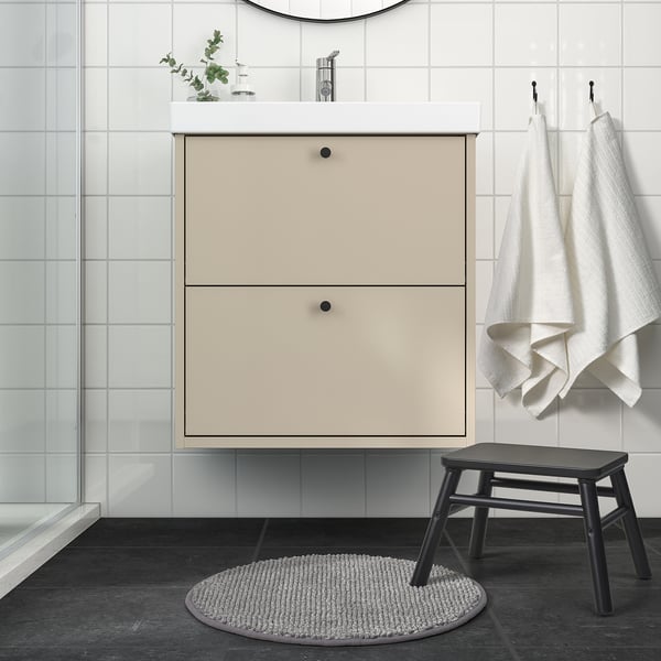 Modernt badrum med ljusbeige skåp, svart pall, mjuk rund TOFTBO-matta vid handfatet, hängande handdukar och duschdörr i glas.