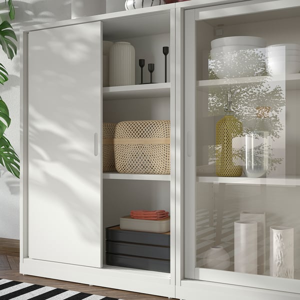Vit förvaringsmöbel TONSTAD med skjutdörrar och glasdörrar, med flätade korgar, vaser och lådor. Modern, benvit design med rena linjer.