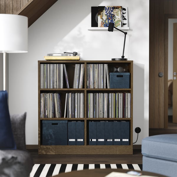 En hylla i TONSTAD i trä med vinylskivor och förvaringslådor. Den är brun, rektangulär och ligger mellan möbler, med en lampa och en stol i närheten.