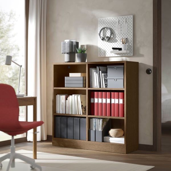 Bokhylla i trä med ljusbrun yta som håller böcker och mappar mot väggen med vit pärlplatta, röd stol och skrivbord i närheten.