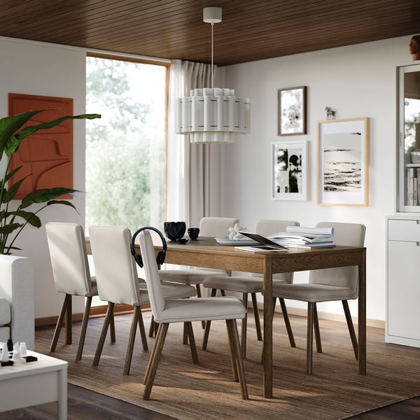 Modern matsal med träbord och 6 stolar, vit inredning, böcker, växt, stort fönster, konst och elegant armatur.