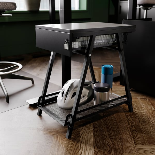 Svart TROTTEN förvaringsbord med låsbar låda och näthylla rymmer olika föremål, placerade på trägolv på kontoret.