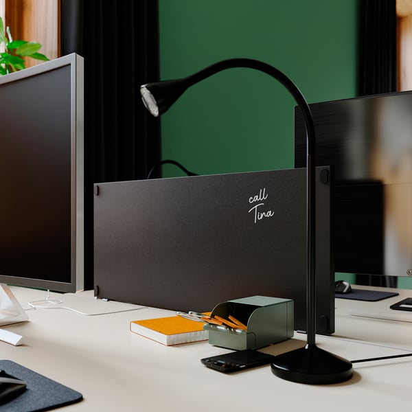 Kontorsarbetsplats med svart trottén skrivbordspartition, lampa, bildskärmar och brevpapper. Grön vägg bakom.