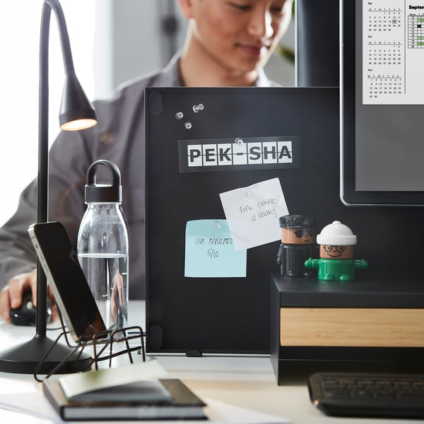 En arbetsyta med en TROTTEN dubbelsidig notis och whiteboard, monterad på ett skrivbord. Den innehåller papper, anteckningar och små dekorationer som hjälper till att organisera organisationen.