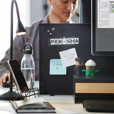 En arbetsyta med en TROTTEN dubbelsidig notis och whiteboard, monterad på ett skrivbord. Den innehåller papper, anteckningar och små dekorationer som hjälper till att organisera organisationen.