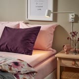 Mysig säng med rosa lakan och inredning, tänd lampa, klocka och blommor.