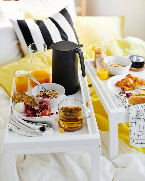 Frukostbricka med vita kuddar; apelsinjuice, croissanter, flingor och en svart kaffetermos.