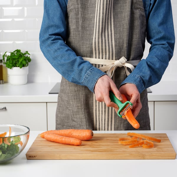 Person som skalar morötter med skalaren UPPFYLLD på skärbräda av trä. Skål med greener i närheten.