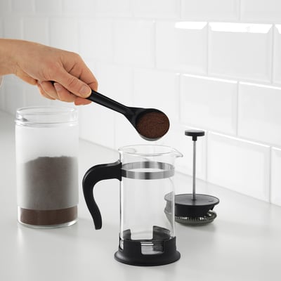 En person tillsätter kaffesump till en klar UPPHETTA fransk press. Kolven är synlig bredvid den, redo att användas på en bänk.