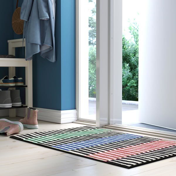 Flerfärgad randig dörrmatta vid skohylla på vitt golv.