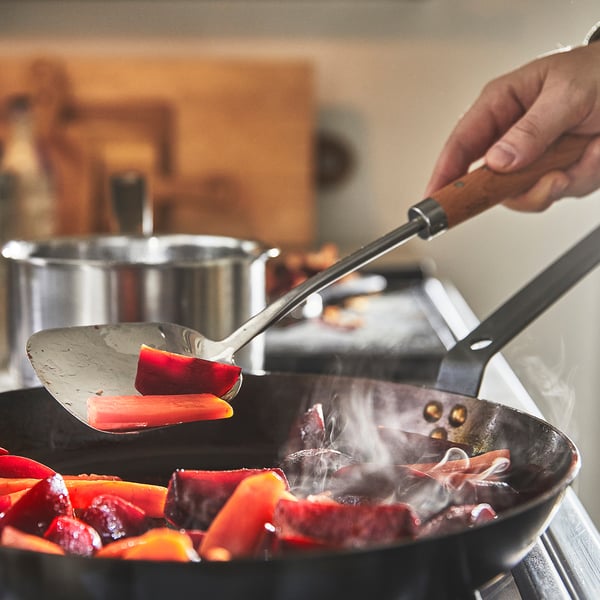 Personen vänder färgglada grönsaker i svart stekpanna, med hjälp av metallspatel. Ånga stiger, vilket visar tillagningsverkan.