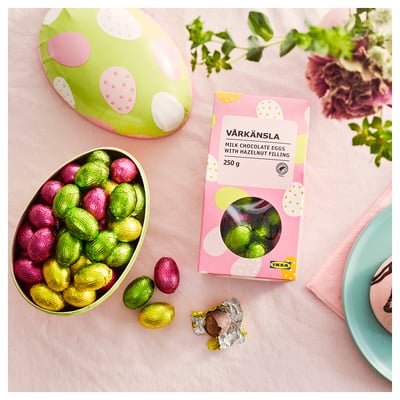 En färgglad folieförpackad mjölkchokladägg i en skål bredvid en låda märkt VÅRKÄNSLA med en rosa, grön och vit design.