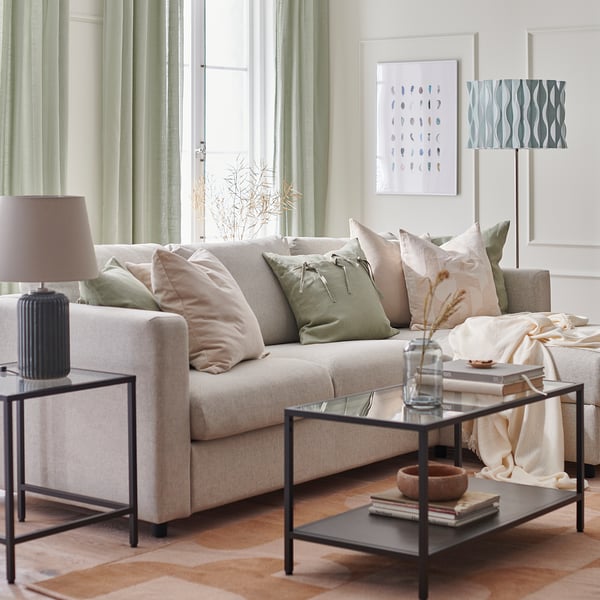 Modernt vardagsrum med gräddvit VIMLE soffa, gröna kuddar, soffbord i glas med böcker, lampa och abstrakt konst.