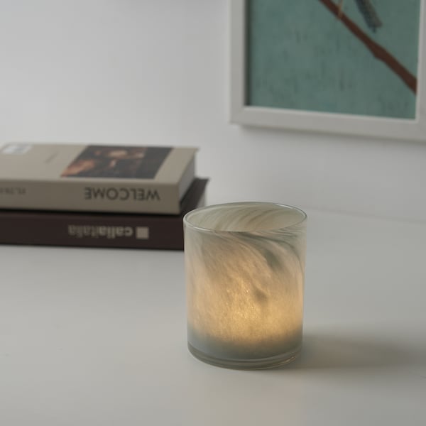 En liten ljusstake i frostat glas står på ett vitt bord och reflekterar ljuset mjukt. Bakom den står böcker staplade på högar.