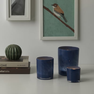 Tre ljusstakar i blått glas med texturerade mönster på bordet, bredvid grön glaskula, under inramad konst.