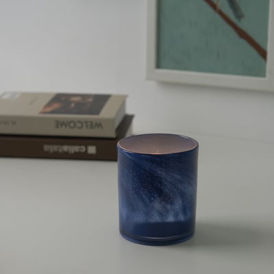 En blå, spräcklig ljusstake står på ett vitt bord, med ett tänt ljus inuti som kastar ett varmt sken. Bakom den syns en bok och en ram.