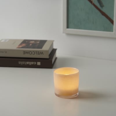 Ett litet tänt ljus står på ett vitt bord bredvid en trave böcker och ger värme och ljus till den fridfulla miljön.