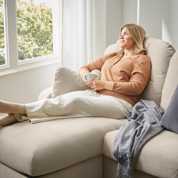 Avkopplande kvinna på beige VINLIDEN soffa, som håller en kopp. Mjuka kuddar, mysig atmosfär vid fönstret.