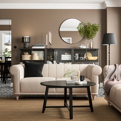 Vardagsrum med soffan VISKAFORS beige, svart soffbord, rund spegel, golvlampa och dekorerade hyllor.