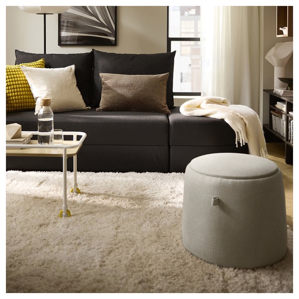 Modernt vardagsrum med svart soffa, vit ryamatta och oval ottoman. Accentuerad med kuddar, ett sidobord och en mysig filt.