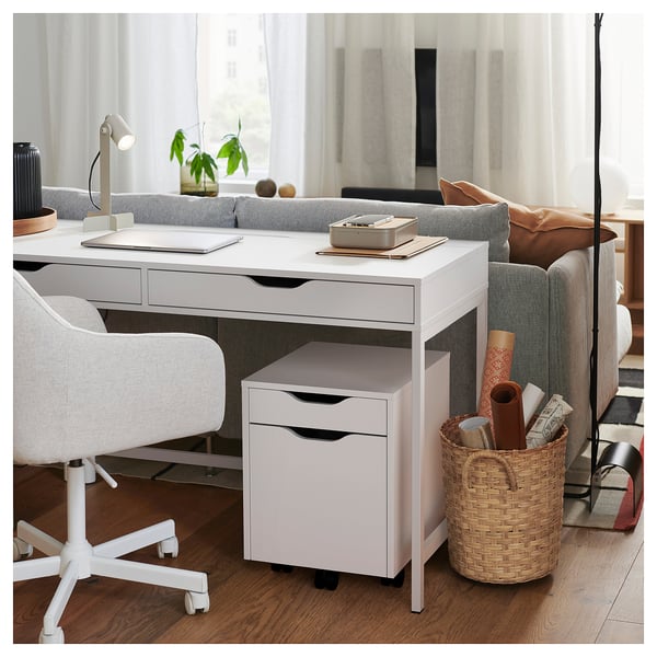 Configuration moderne du bureau à domicile avec un bureau et une chaise ALEX blancs, comprenant un ordinateur portable, un panier avec des fournitures et un canapé douillet avec un coussin et une couverture.