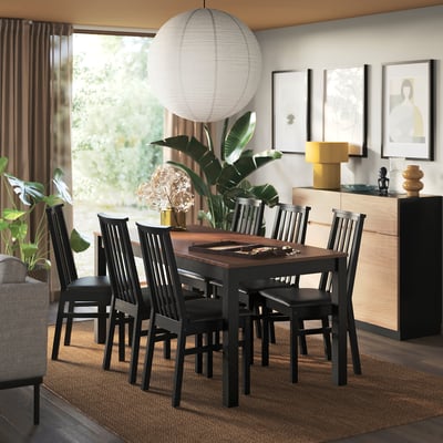 Une salle à manger moderne avec une chaise ÅLHULT sombre assise autour d’une table en bois, complétant la décoration contemporaine environnante et de grandes fenêtres.