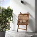 Chaise en bois sur le balcon, appuyée contre le mur, à côté d’une plante en pot.