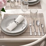 Table avec napperon gris, vaisselle blanche, serviette roulée, verrerie et couverts polis, y compris une fourchette, un couteau et une cuillère.